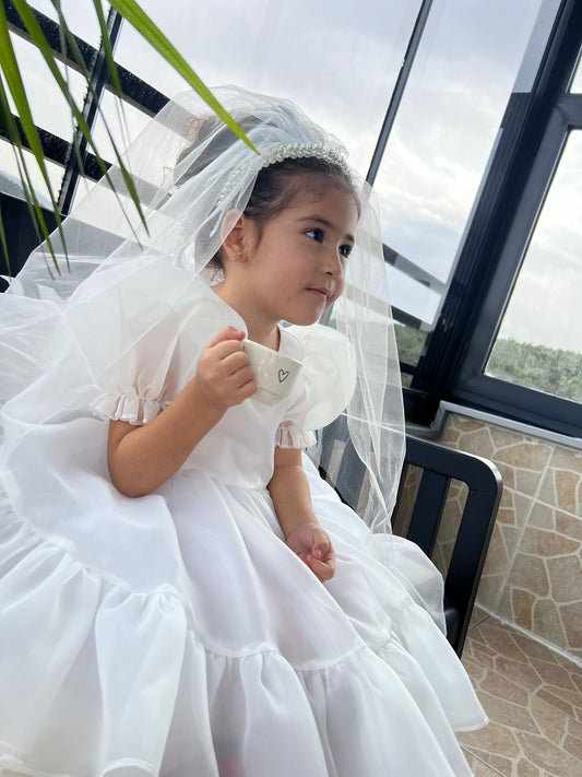 Girl in white organza communion dress holding tulle skirt in a cozy indoor setting