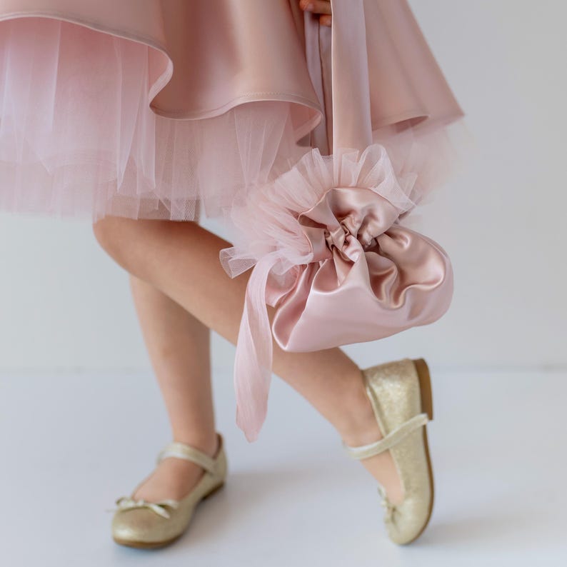 "Close-up of a pink flower girl dress with a tulle skirt, paired with a satin handbag and gold shoes.