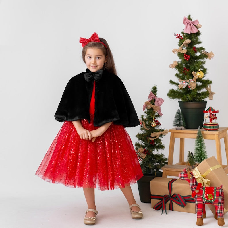 Girl wearing a red satin flower girl dress styled with a black cape, shown as a sparkly Christmas holiday outfit.
