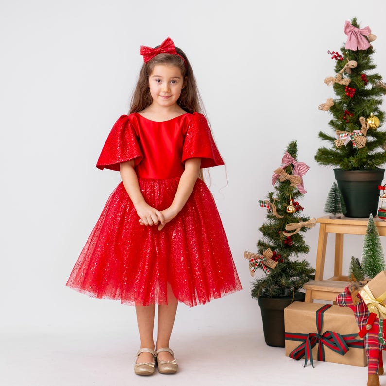 Red satin flower girl dress with sparkly tulle skirt and short flutter sleeves, shown in a Christmas studio setting.