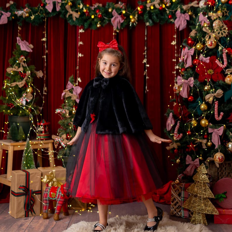 Girl wearing a red and black sparkly Christmas holiday dress styled with a black faux fur cape in a festive decorated setting.”