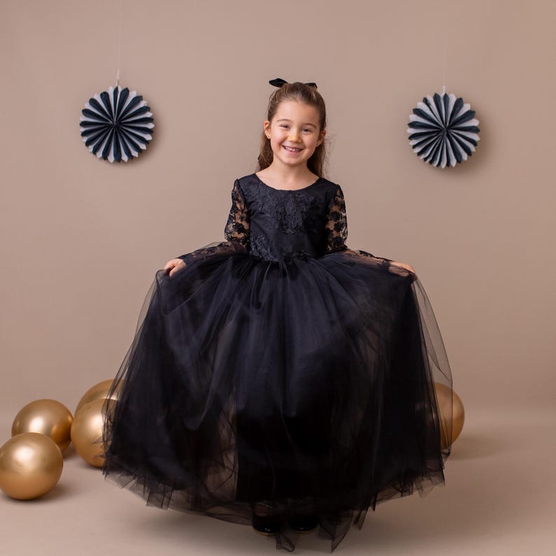 Little girl in a black lace tulle flower-girl dress, standing with gold balloons and decorative wall elements.