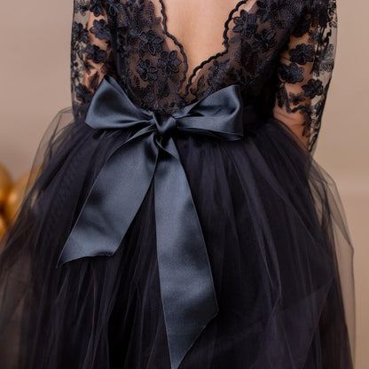 Young girl in a black lace dress with a large black bow, set against a neutral beige background.