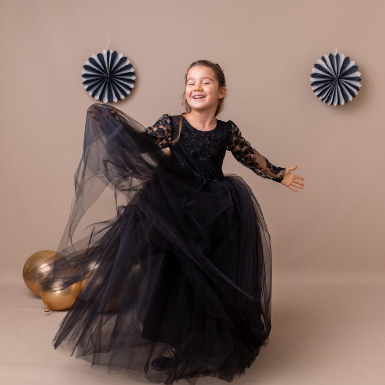 Little girl twirling in a black lace long-sleeve flower girl dress with a layered tulle skirt, studio background.
