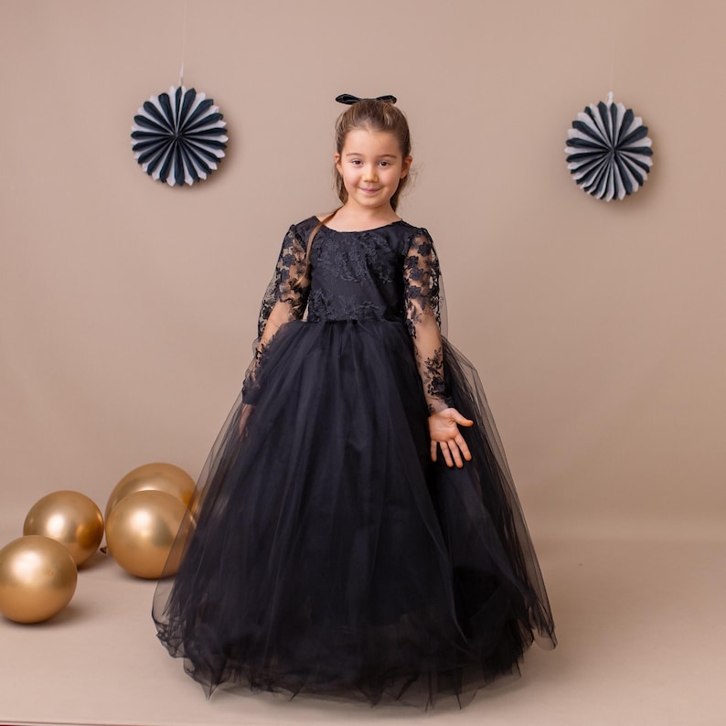 Black lace long-sleeve tulle flower girl dress displayed on a young model standing against a beige backdrop.