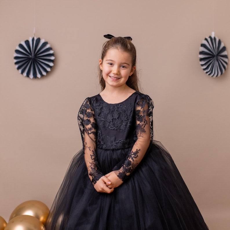 Black lace long-sleeve tulle flower girl dress worn by a young girl standing against a beige decorated background.