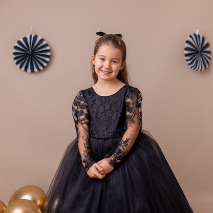 Black lace long-sleeve tulle flower girl dress worn by a young girl standing against a beige decorated background.