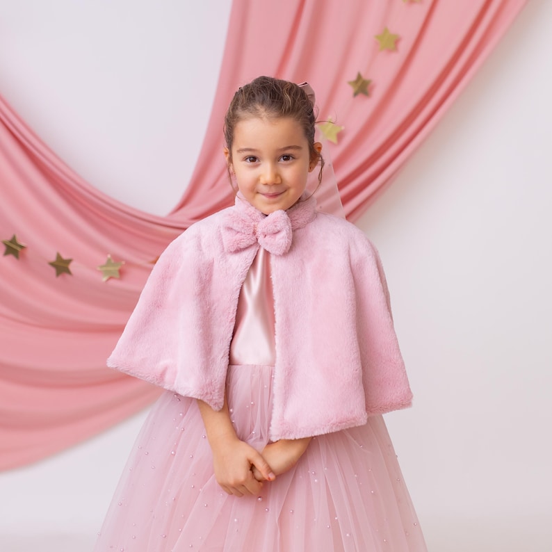 Young girl in a pink flower girl dress with a faux fur cape, featuring a bow detail, standing with a soft pink backdrop.