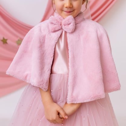 Close-up of a young girl in a pink flower girl dress with a faux fur cape and a bow detail, standing with a soft pink backdrop.