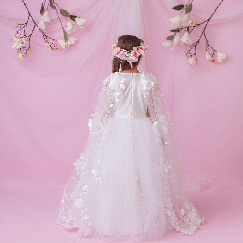 Back view of a young girl in a white personalized flower girl dress with a floral cape and crown, against a soft pink backdrop.