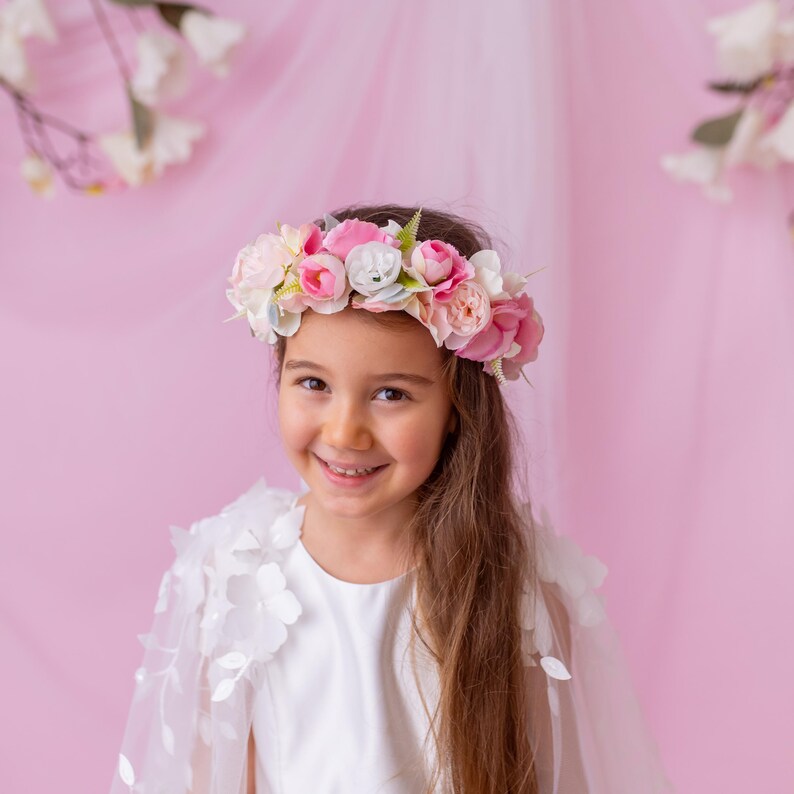 Close-up of a young girl in a white flower girl dress with a floral cape, wearing a vibrant flower crown and smiling.