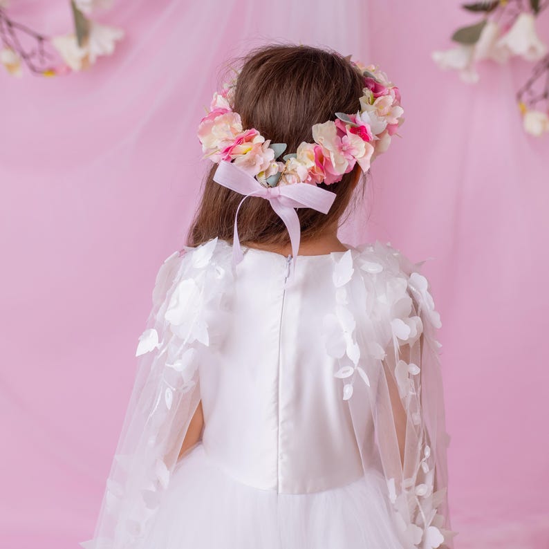 Back view of a young girl in a white flower girl dress with a floral cape, wearing a flower crown tied with a ribbon.