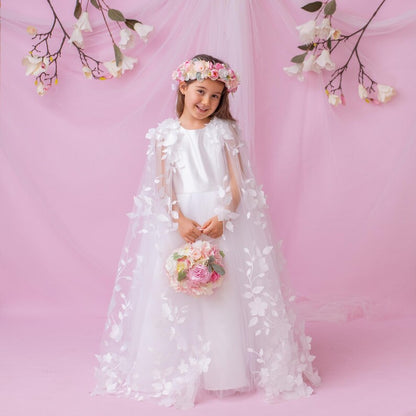Young girl in a white flower girl dress with a floral cape and crown, holding a flower bouquet, smiling against a pink backdrop.