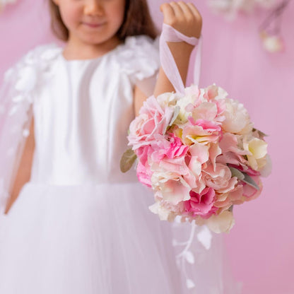 Close-up of a young girl in a white flower girl dress with a floral cape, holding a bouquet of pink flowers.