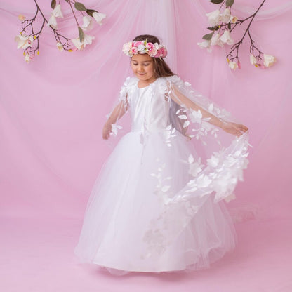 Young girl in a white personalized flower girl dress with a floral cape, twirling and smiling against a soft pink backdrop.