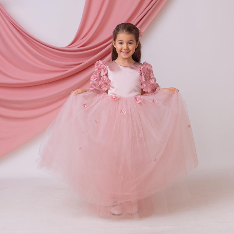 Front view of blush satin flower girl dress holding the tulle skirt on both sides, highlighting volume and floral embellishments