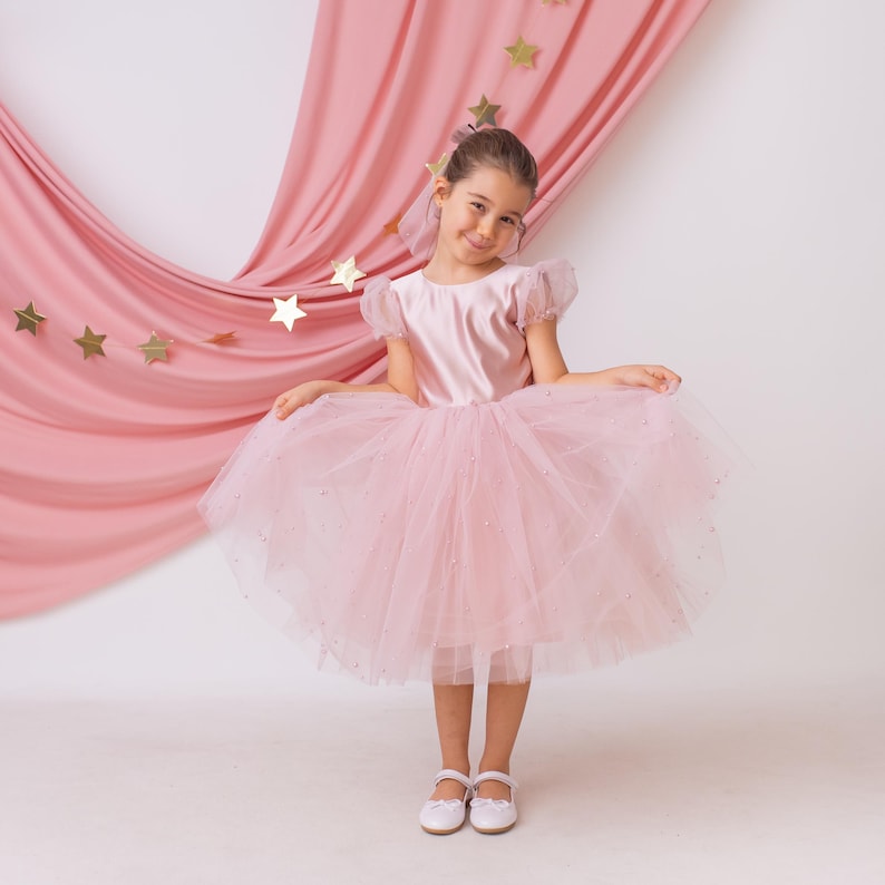 Girl in a pink flower girl dress with a tulle skirt, holding the skirt playfully with golden stars in the background.