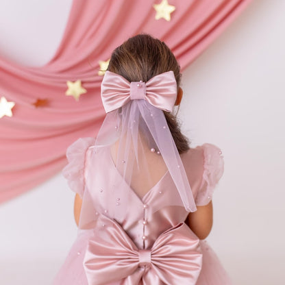 Back view of a pink flower girl dress with a satin bow, veil, and pearl details, with a soft pink backdrop.