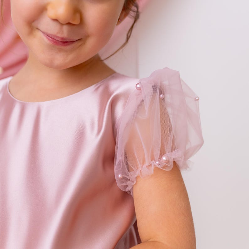 Close-up of a pink flower girl dress with sheer puffed sleeves and pearl details, with a soft pink backdrop.