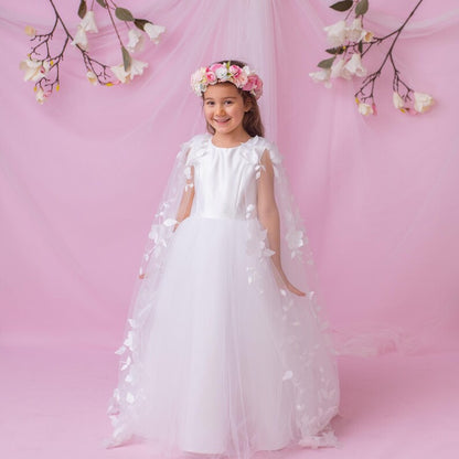 Young girl in a white personalized flower girl dress with a floral cape and crown, posing against a soft pink backdrop.