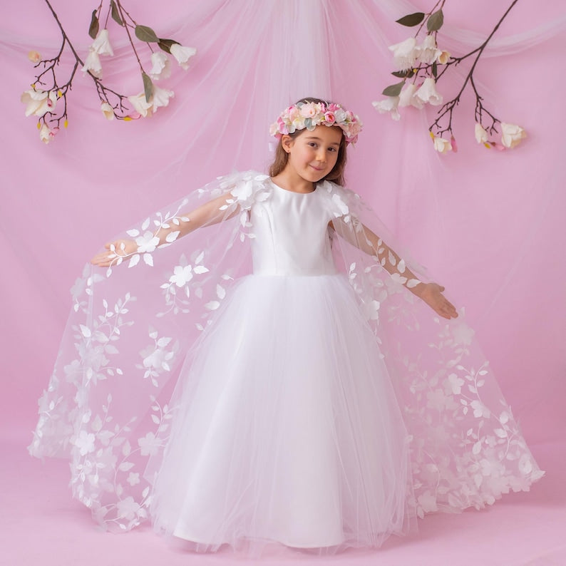 Young girl in a personalized flower girl dress with floral cape, wearing a flower crown, set against a pink backdrop.